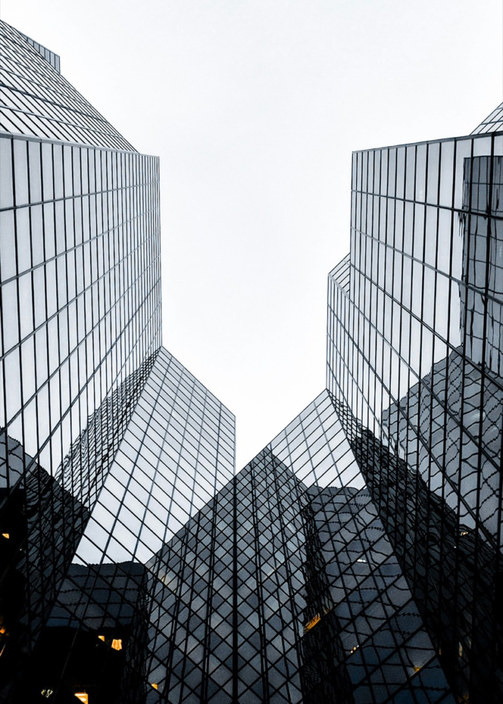 Two glass skyscrapers rising toward each other against a gray sky, viewed from below